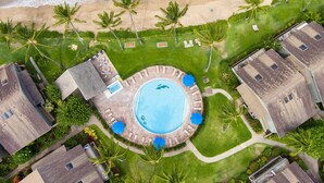 Eine Vogelperspektive zeigt einen Urlaubsort mit einem zentralen Schwimmbad, Zugang zum Strand und mehreren bungalowartigen Gebäuden.