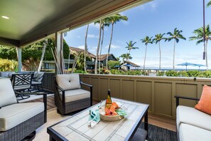 Een overdekt terras met rieten meubels, een tafel met een fles en bloemen, en een uitzicht op palmbomen en een gebouw op de achtergrond.