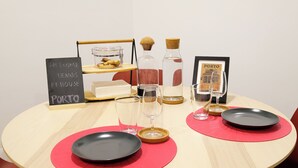 Une table ronde est dressée avec des assiettes noires, des sets de table rouges et des verres à vin transparents. Un panneau noir affiche l’inscription : « BIENVENUE CHEZ DENNIS FE HOUSE PORTO ».