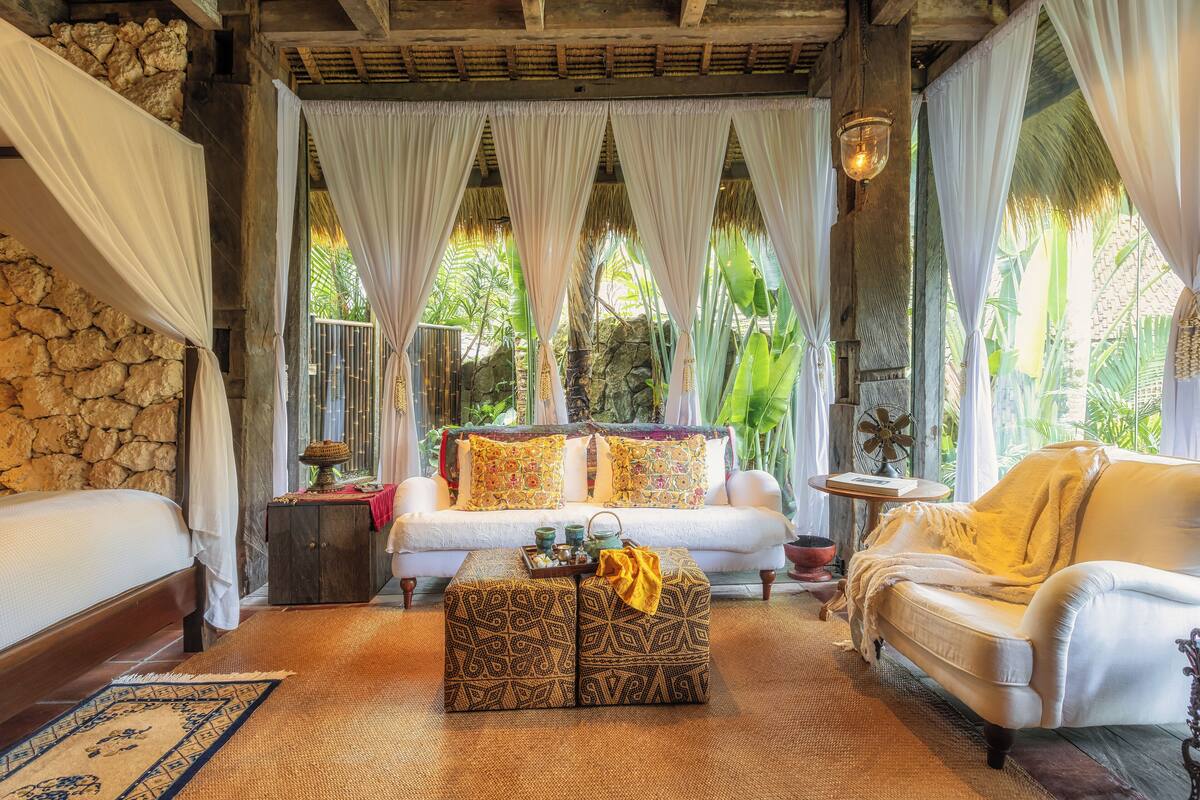 A cozy room with a bed, a sofa, and a patterned ottoman. There is a small table with a lamp and a decorative item. The room has a stone wall and large windows with curtains.