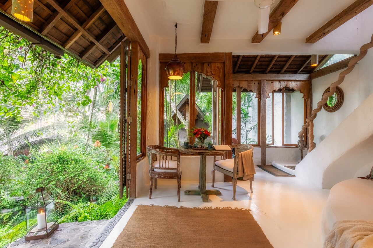 A room with a wooden ceiling, large windows, a dining table with chairs, and a view of lush greenery outside.