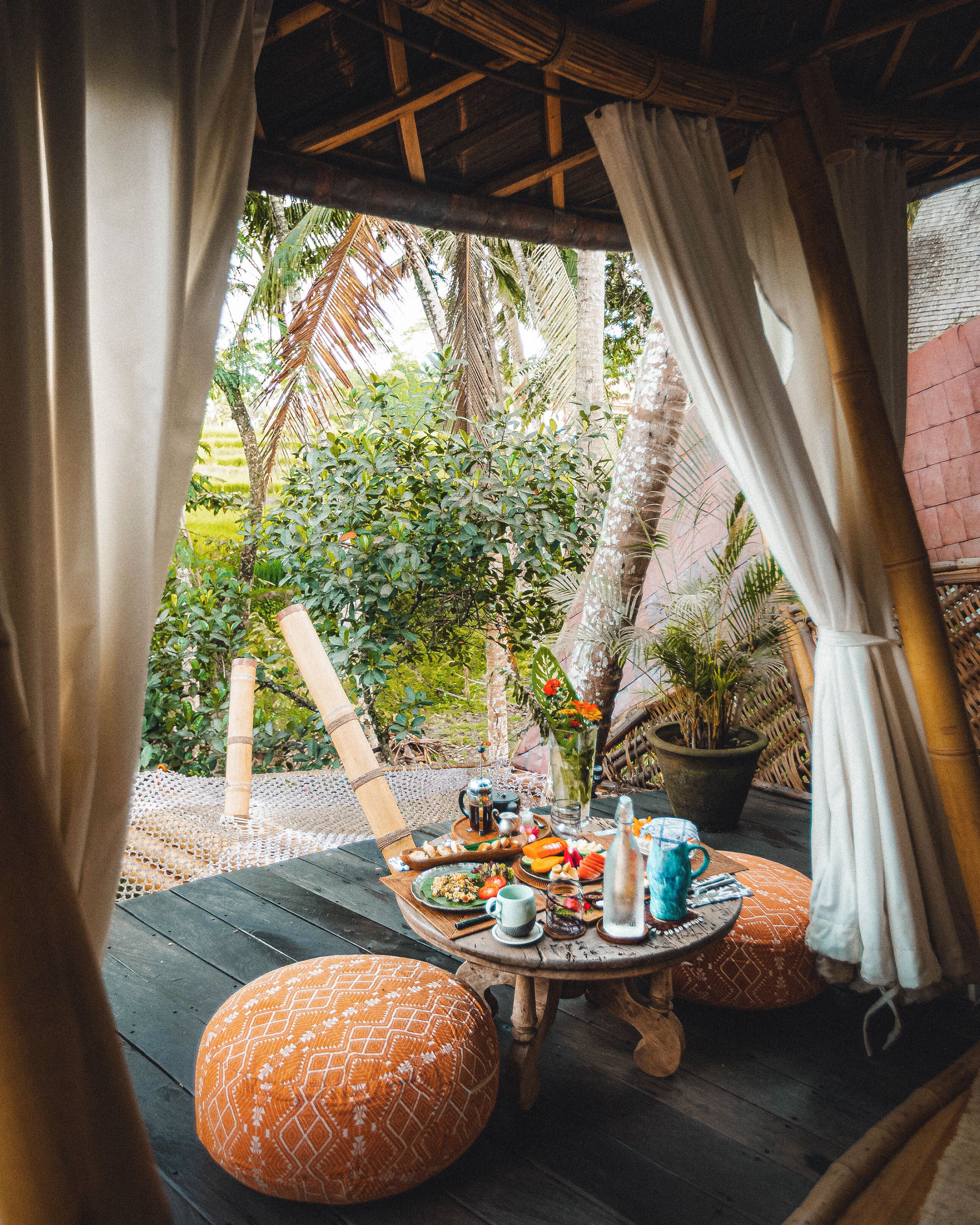 An outdoor dining area with a round table set for a meal, surrounded by cushioned seating, and a view of tropical vegetation.