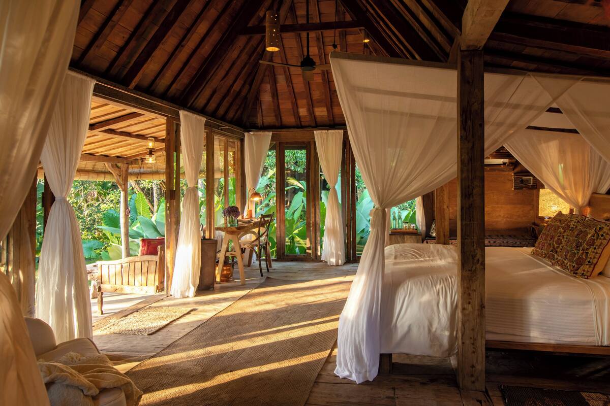 A room with a wooden ceiling, a bed with a canopy, a table with chairs, and a view of greenery outside.
