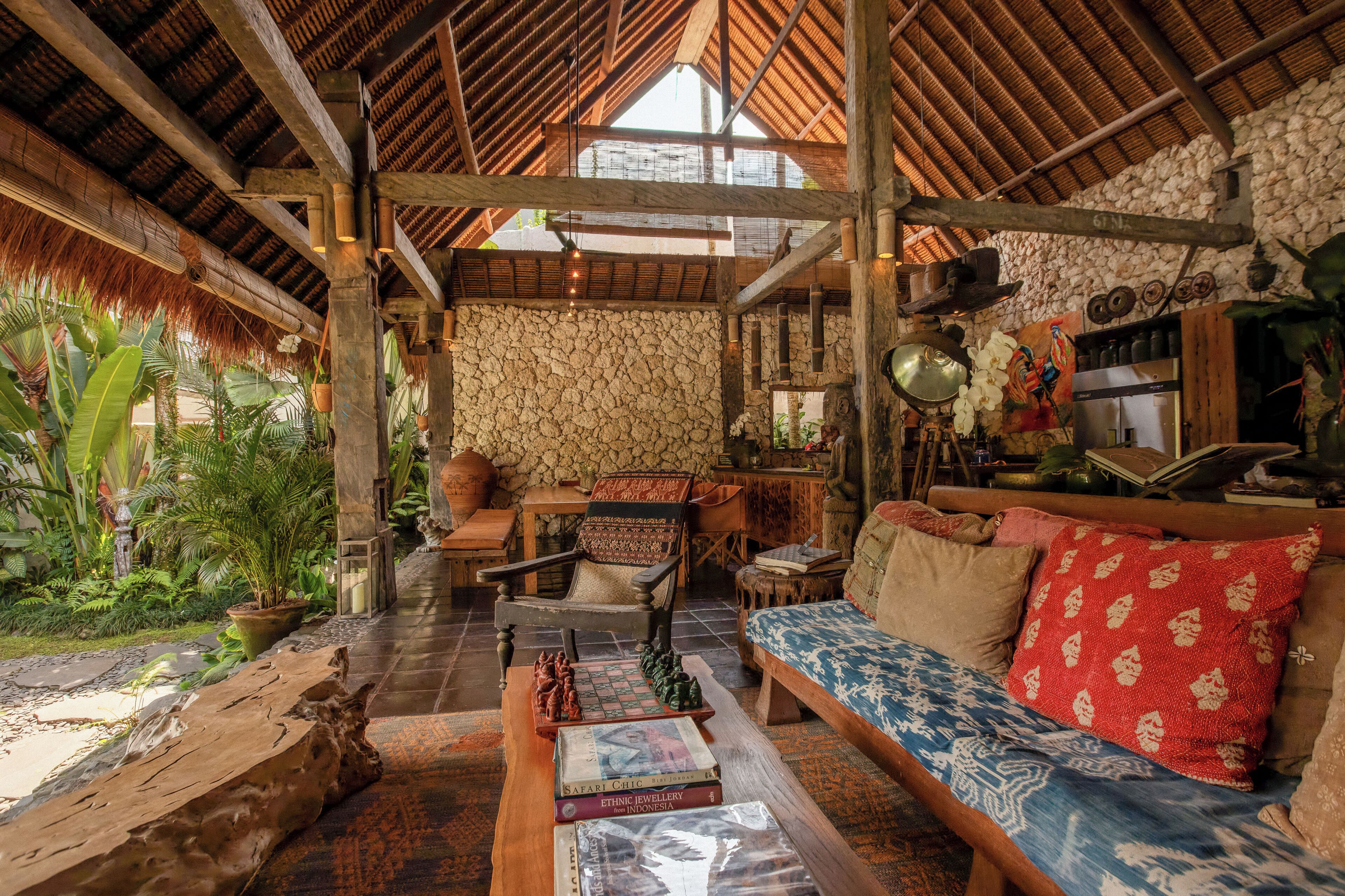 A spacious room with a high ceiling, a stone wall, and a thatched roof. There is a wooden bench, a sofa with patterned cushions, and a table with books and decorative items.