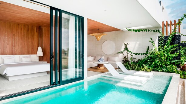 Un área de piscina interior moderna con una puerta de vidrio que da a un dormitorio, un sofá blanco y un sillón.