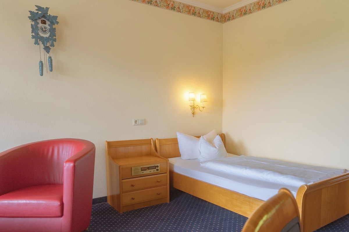 A hotel room with a single bed, a wooden nightstand, a red chair, and a wall clock.