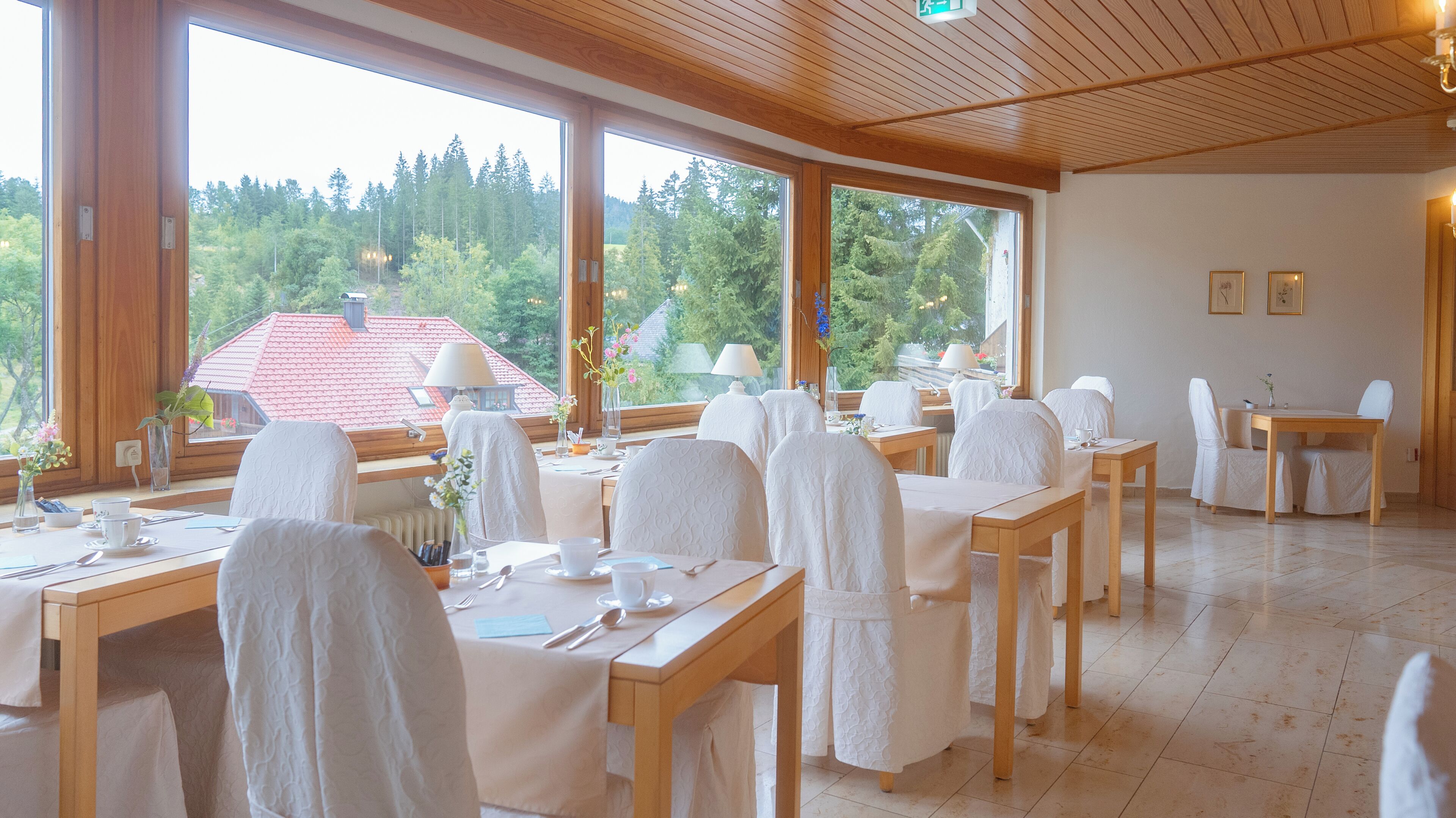 A dining area with tables set for two, large windows offering a view of a forested area, and a wooden ceiling.