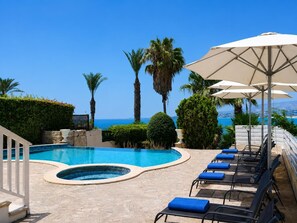 A pool area with a hot tub, lounge chairs, and umbrellas, overlooking a clear blue sea.