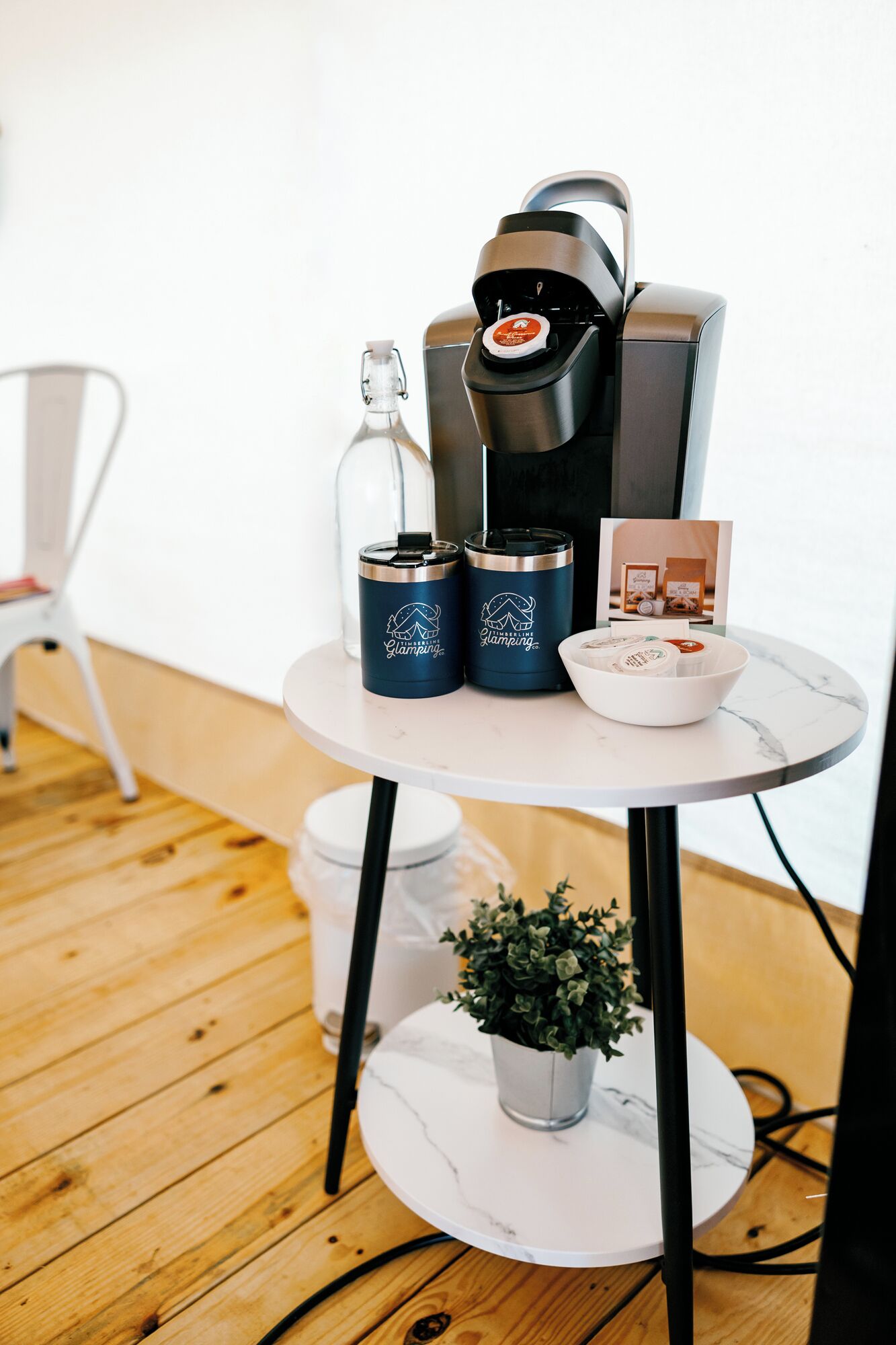 Een tafel met een koffiezetapparaat, twee thermosflessen, een glazen fles, een kleine plant en een schaal met items, alles op een witte tafel met een marmeren blad.