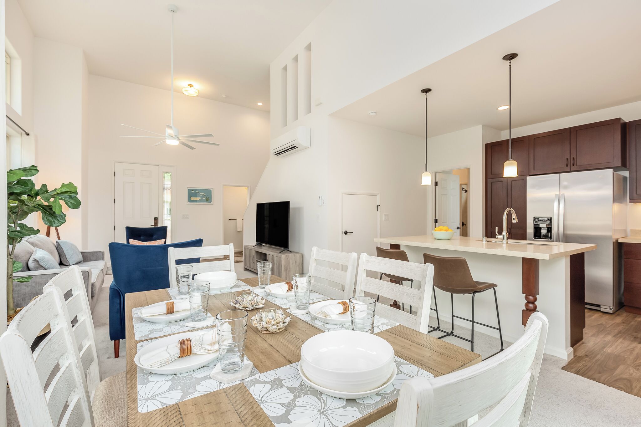 A modern kitchen with a central island, a dining area with white chairs and a wooden table, and a living room with a sofa and a TV.