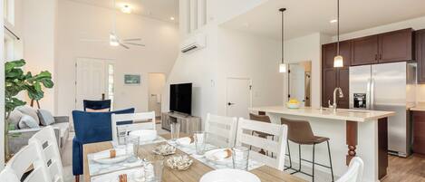 A modern kitchen with a central island, a dining area with white chairs and a wooden table, and a living room with a sofa and a TV.