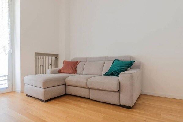 A light-colored sectional sofa with two cushions, a radiator, and a wooden floor in a room with white walls.