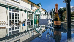 A table with a bottle of Canti Prosecco DOC and two wine glasses, reflecting a building with green trim and a clear blue sky in the background.