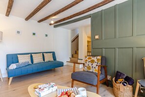 Un saló amb un sofà blau, una cadira de fusta, una taula amb llibres i fruita, i una escala al fons.