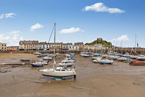 Une ville côtière bordée d’une rangée de bâtiments, une colline surmontée d’un château et de nombreux bateaux amarrés sur la plage.