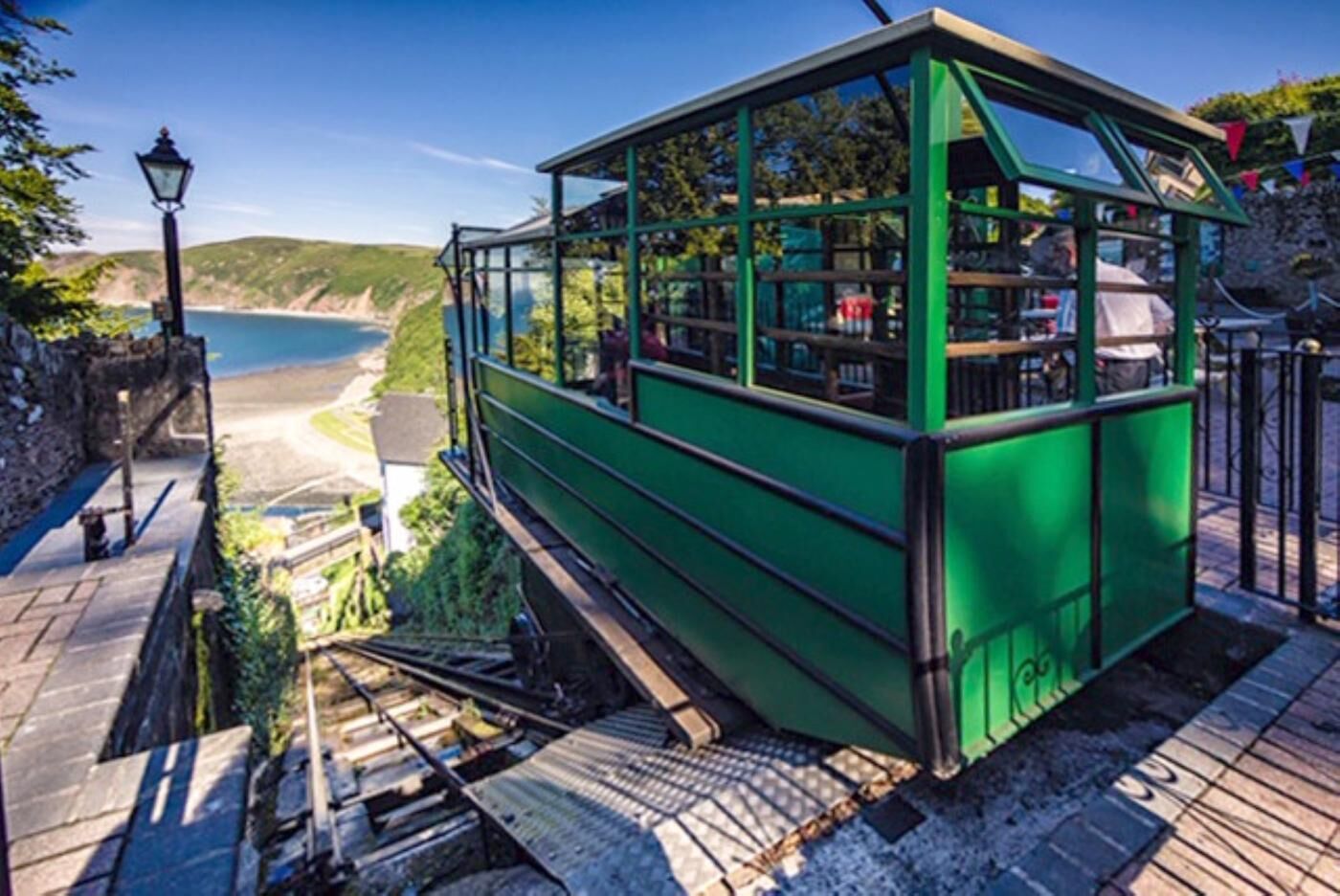 Une télécabine verte avec une structure en verre, une plateforme en bois et un mur en pierre.