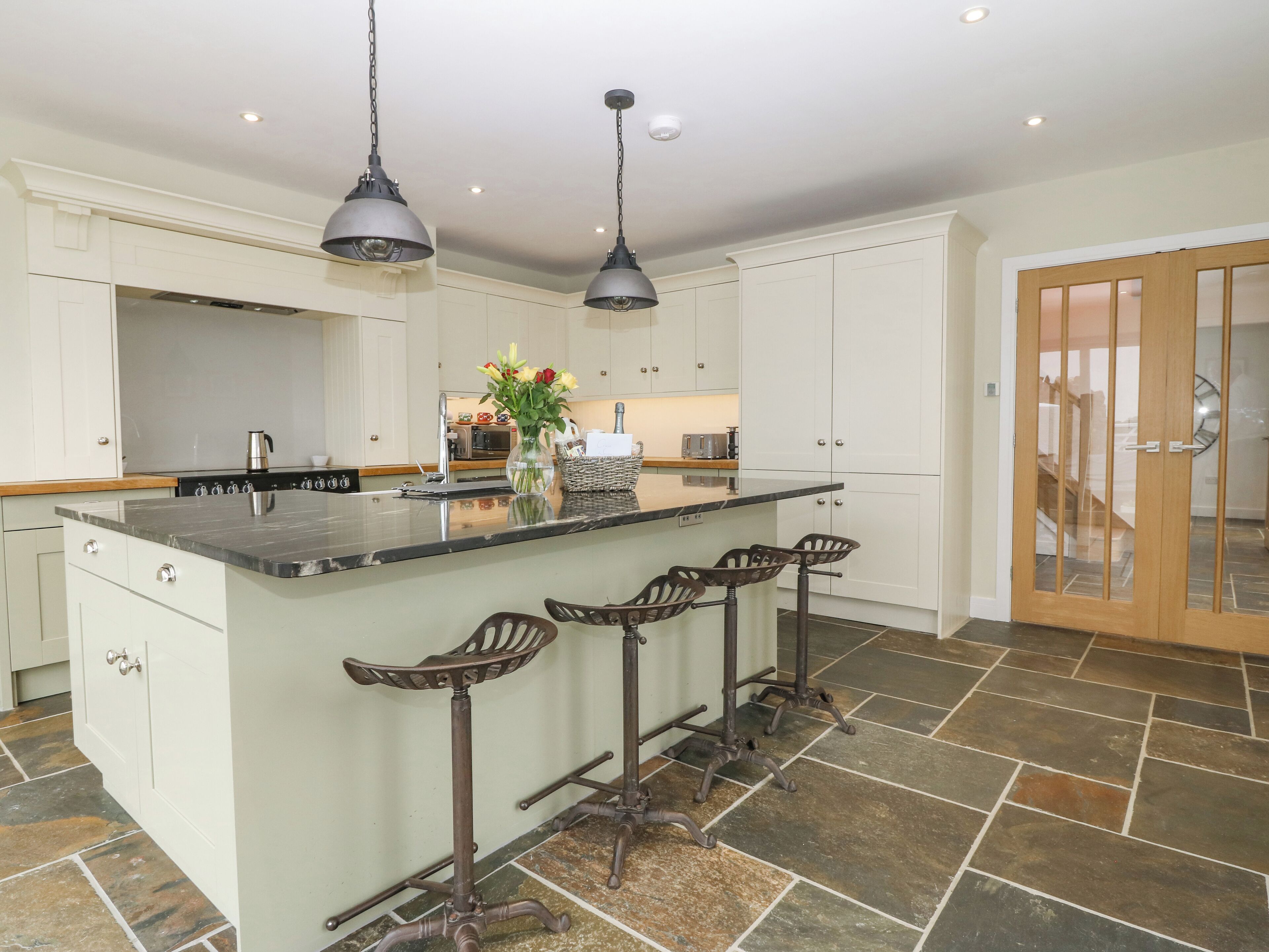 A spacious kitchen with a large island, bar stools, pendant lighting, and a view of the living area through a glass door.