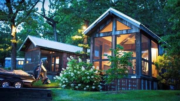 Une cabane dotée d’une grande fenêtre en verre, entourée d’arbres et abritant un véhicule.