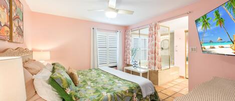 Une chambre à coucher avec un lit, une table de chevet, une fenêtre avec des volets, un ventilateur de plafond et une vue sur la plage, visible à travers un miroir.