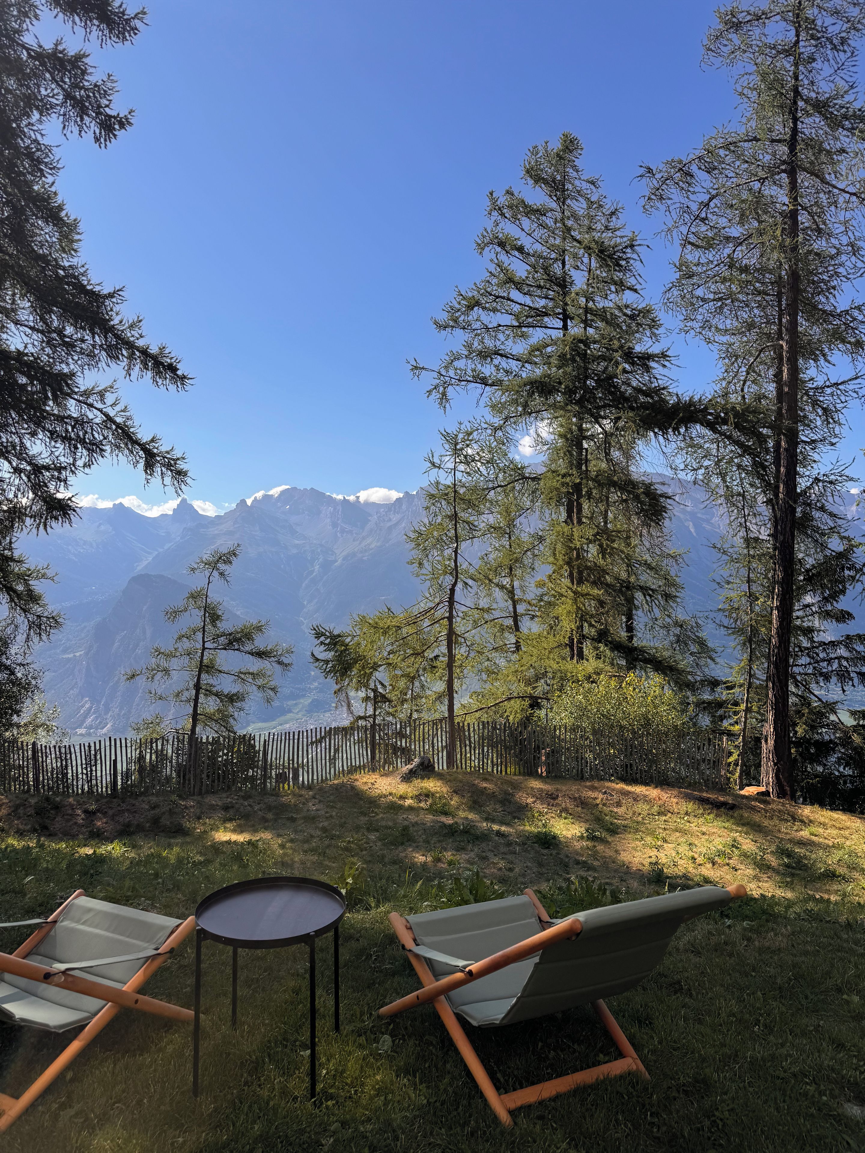 A grassy outdoor area with two lounge chairs and a small table, surrounded by tall trees and a wooden fence, overlooking a mountain range.