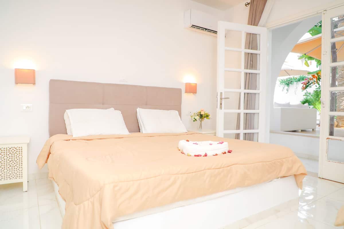Una cama bien hecha con una colcha color melocotón y almohadas blancas, un pequeño jarrón con flores en una mesita de noche, y una puerta blanca que da a un área exterior con plantas.