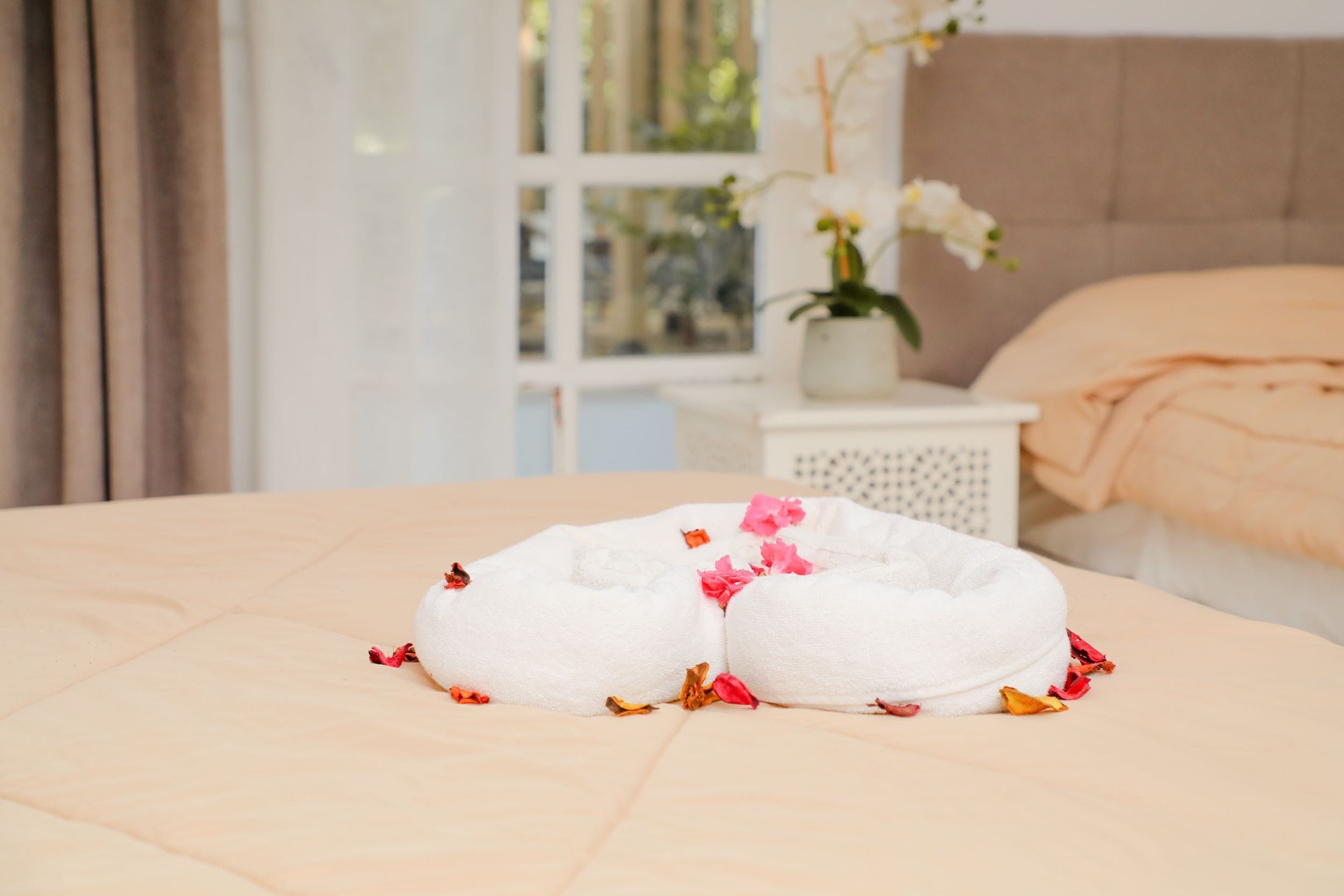 Een net opgemaakt bed met een witte dekbedovertrek en twee opgerolde handdoeken, verspreid met rozenblaadjes.