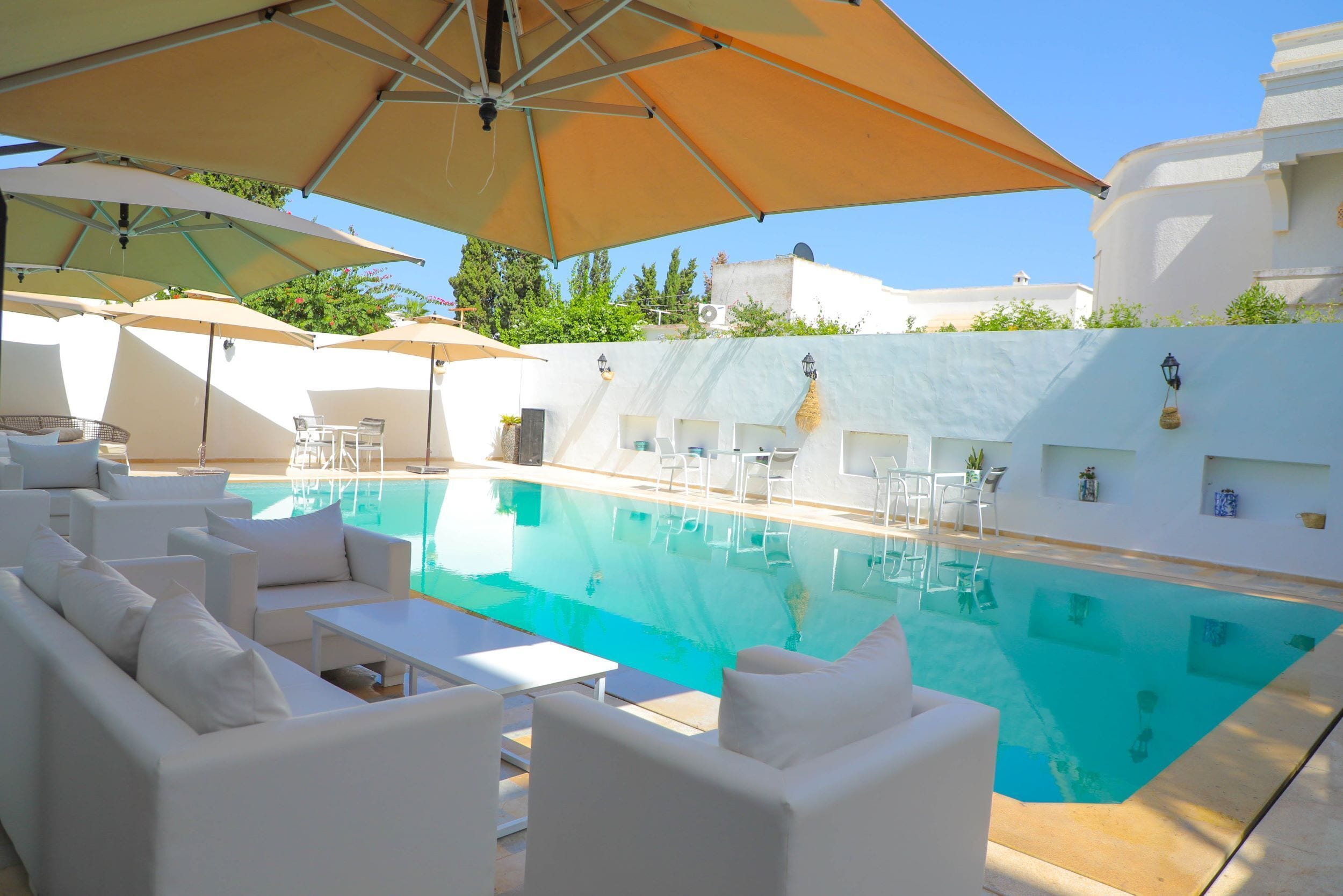 Un espace aménagé au bord de la piscine, avec des chaises longues blanches et des parasols, une piscine à l’eau claire et un bâtiment blanc doté de balcons.