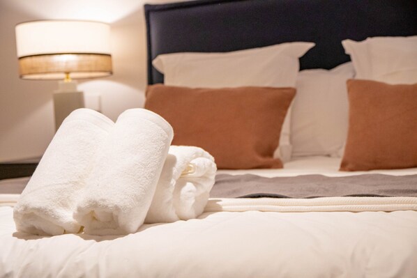 A neatly made bed with white and brown pillows, a folded white towel, and a bedside lamp.