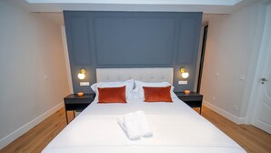 A neatly made bed with white linens, two red pillows, and a folded towel in the center. There are two bedside tables with lamps on either side of the bed.
