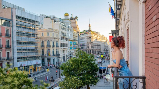 A balcony overlooking a city street with buildings, trees, and pedestrians.