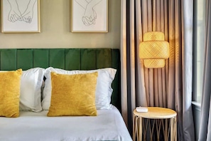 A hotel room with a green upholstered headboard, a bed with white and mustard pillows, a small round table, and a wall-mounted lamp.