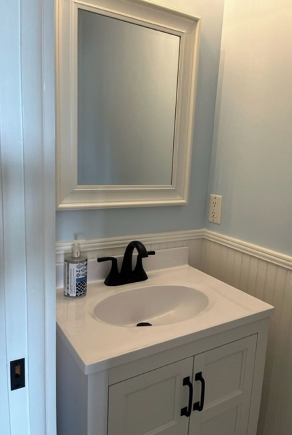A bathroom with a white sink, black faucet, and a mirror above it. There is a bottle of hand soap on the sink counter.