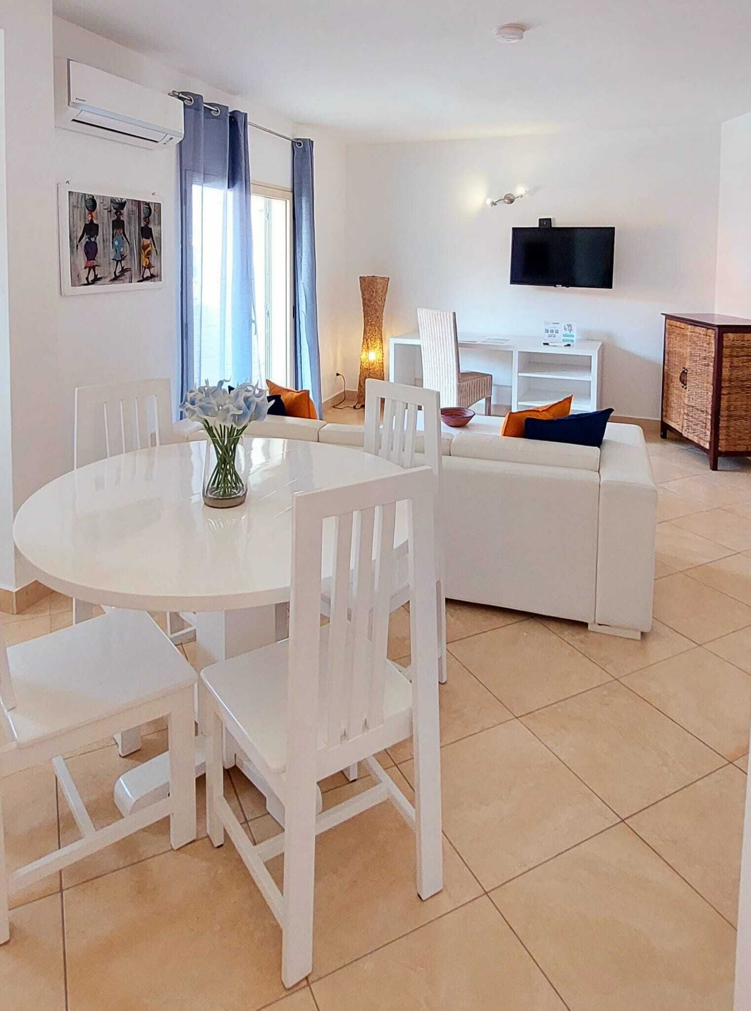 A modern living room with a white round table, white chairs, a white sofa, and a flat-screen TV mounted on the wall.