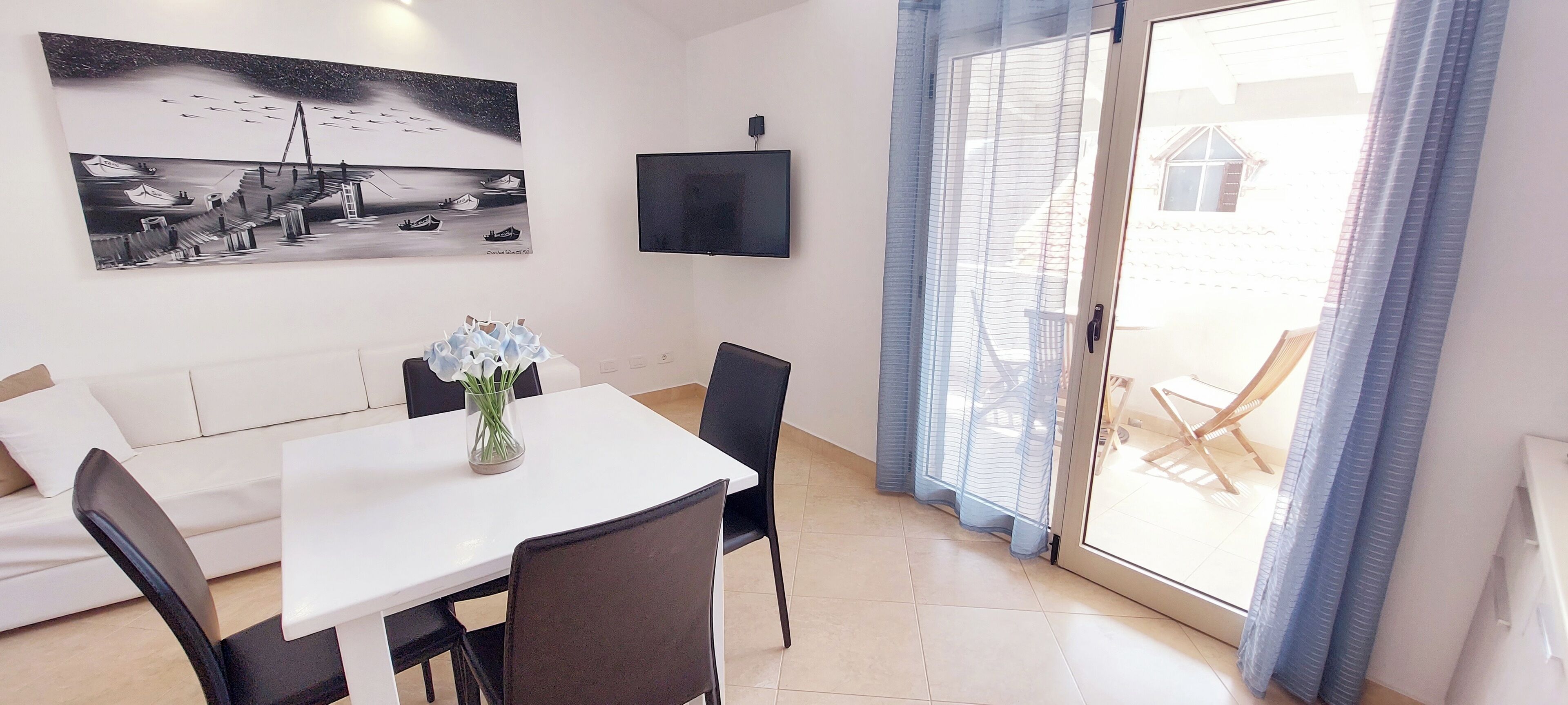 Una sala de estar moderna con un sofá blanco, una mesa de comedor con sillas, un televisor de pantalla plana montado en la pared y un amplio ventanal con cortinas azules.