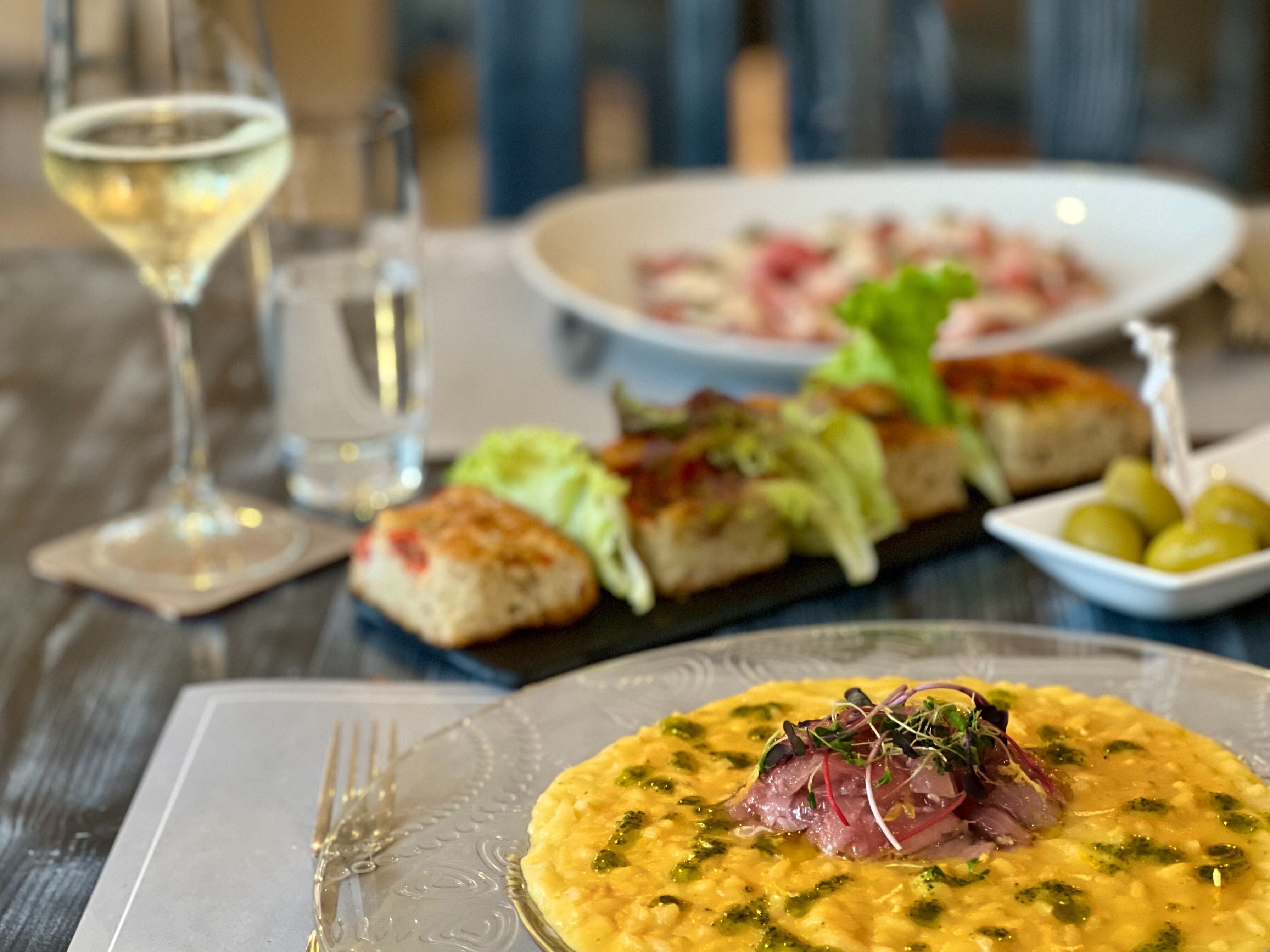 A dining table with a variety of dishes, including a plate of risotto with greens and a piece of meat, a glass of white wine, and a bowl of olives.