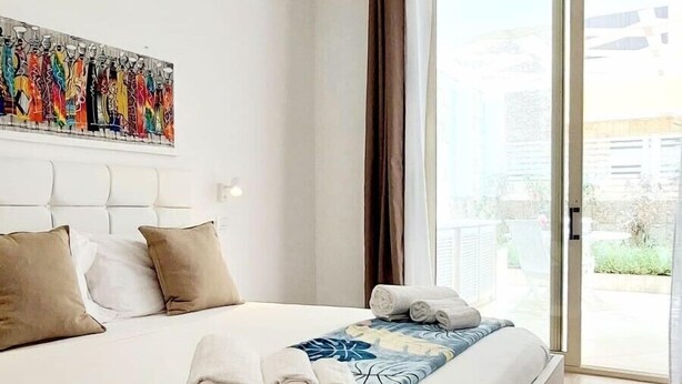 A modern hotel room with a large bed, a view through a sliding glass door, and a colorful abstract painting on the wall. - Branco Suites (Sal)