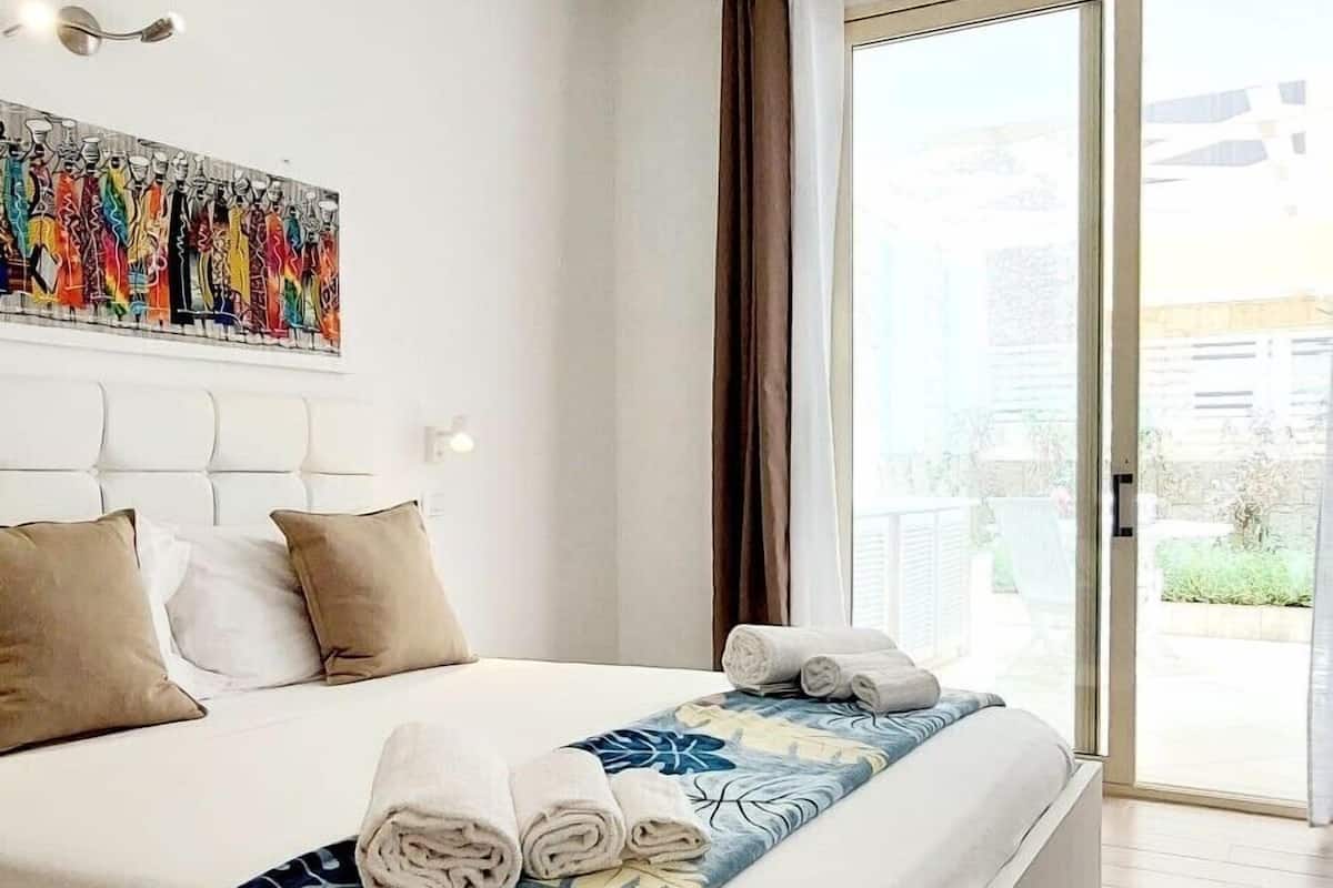 Quarto de hotel moderno com uma cama grande, vista através de uma porta de correr de vidro e uma pintura abstrata colorida na parede.