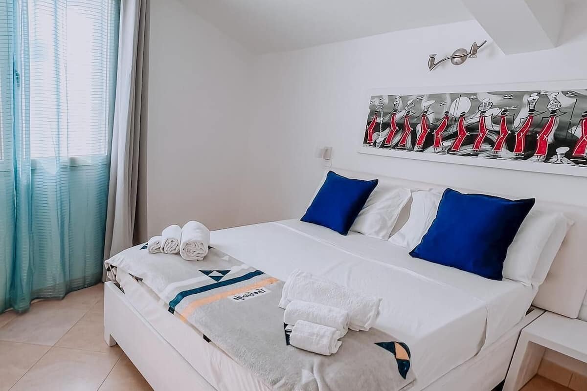 Uma cama bem arrumada com lençóis brancos e almofadas azuis, um tapete com padrão azul e branco, e uma obra de arte na parede retratando chefs.