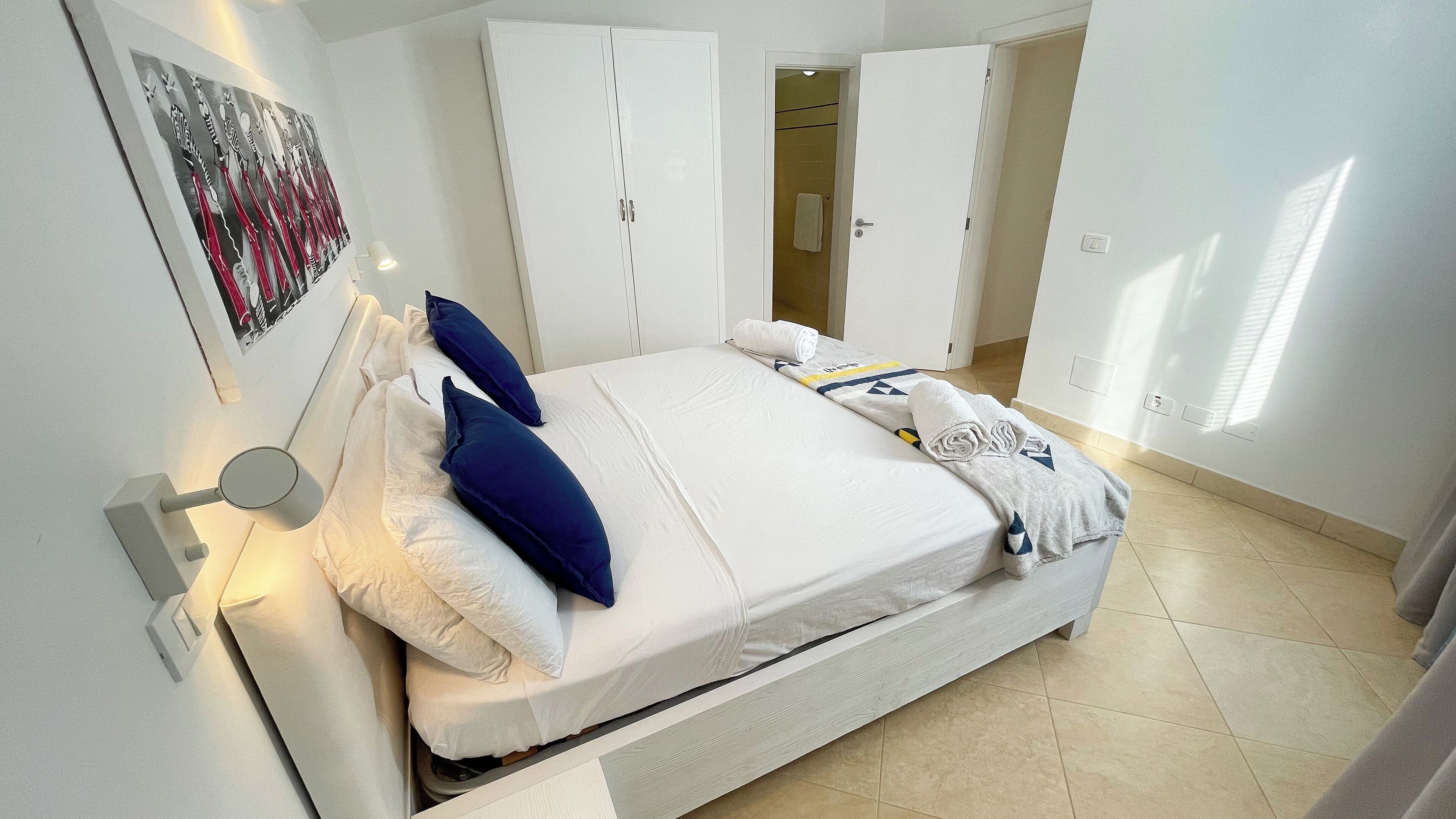 Un lit bien fait, avec des oreillers blancs et bleus, une couverture grise et une serviette enroulée. On y trouve des armoires blanches et une porte qui donne sur une salle de bain.
