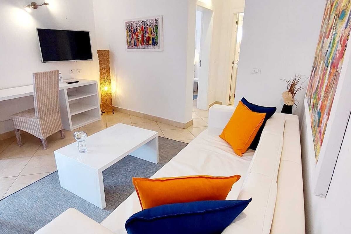 Uma sala de estar moderna com um sofá branco, almofadas laranja e azuis, uma mesa de centro branca, uma TV na parede e um quadro.