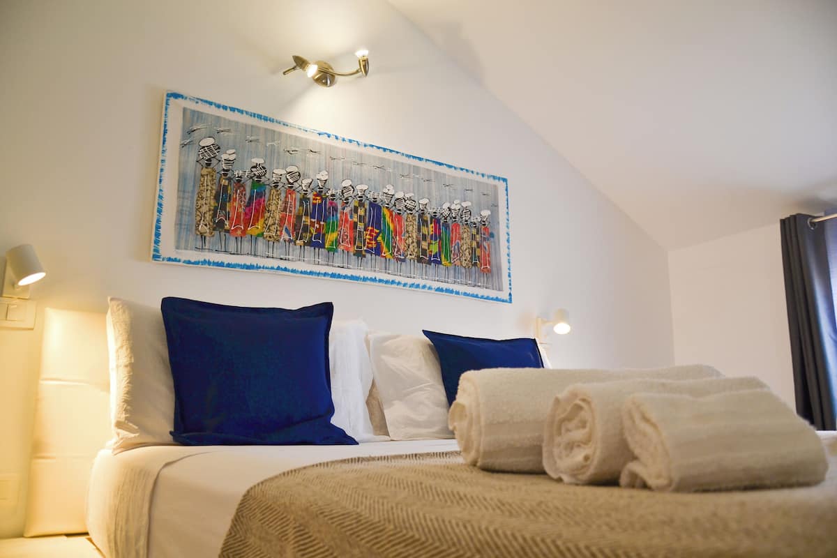 Quarto de hotel com cama, almofadas, uma obra de arte na parede e luminária no teto.