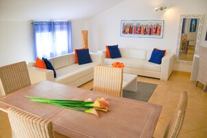 A living room with a dining table, two white sofas with blue and orange cushions, wicker chairs, a large mirror, and a wall art piece.