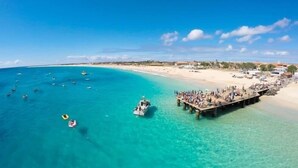 A beach with a pier extending into the water, boats, and a clear blue sky. - Branco Suites (Sal)