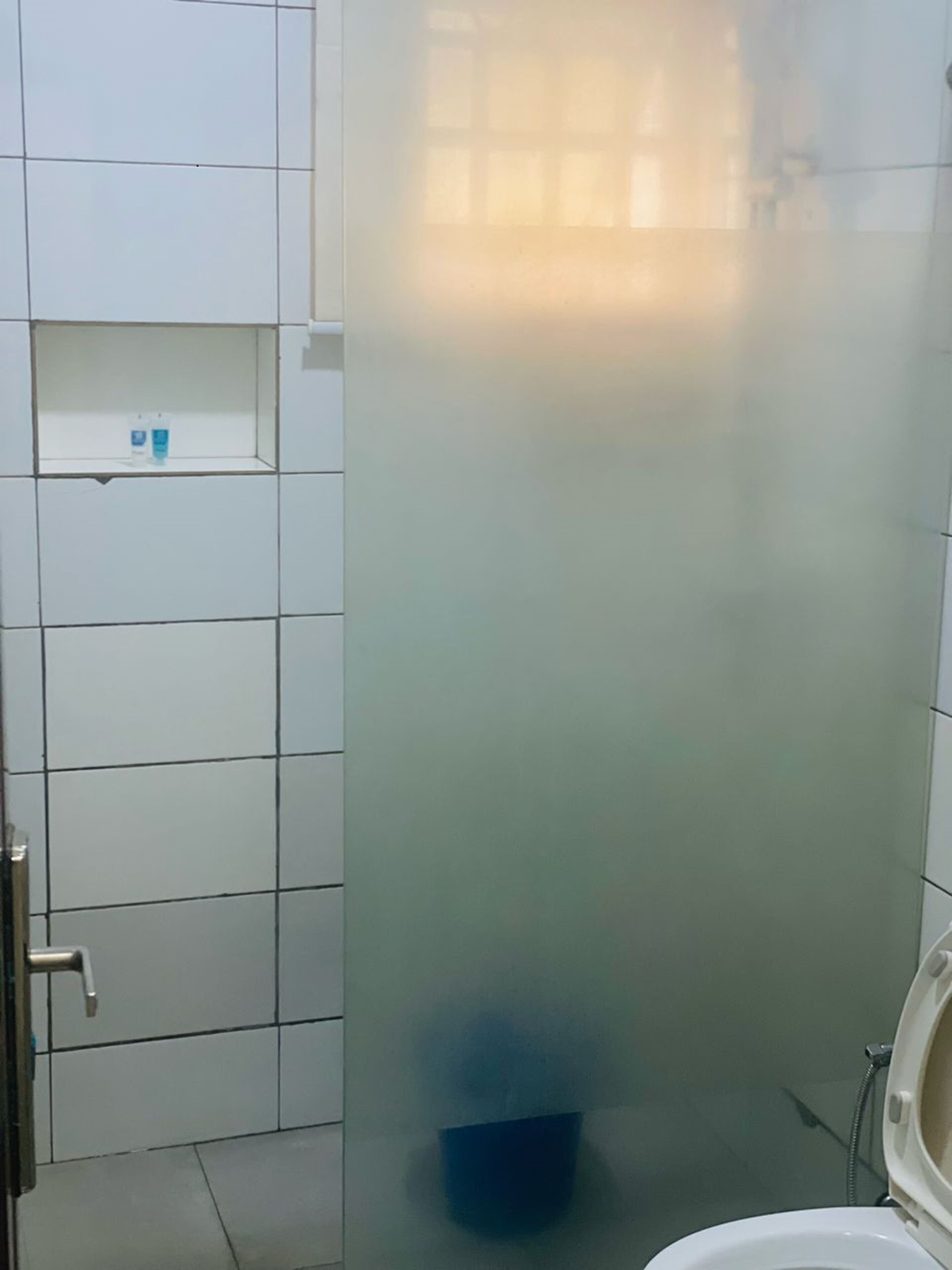 A bathroom with white tiled walls, a toilet, a blue trash bin, and a small shelf with toiletries.