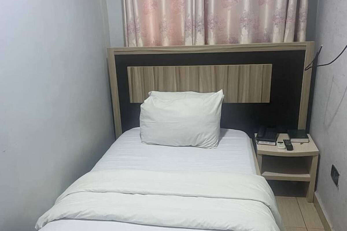 A small, neatly made bed with white linens in a compact room with a wooden headboard and a nightstand with a phone and remote control.
