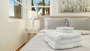 Een net opgemaakt bed met een gewatteerde deken en opgevouwen handdoeken, een nachtkastje met een lamp, en een schilderij aan de muur.