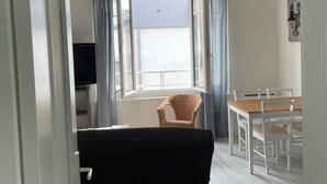 A living room with a black sofa, a dining table with chairs, a wicker chair, and a TV. - L'escale, Appartement au Cœur de Granville (Granville)