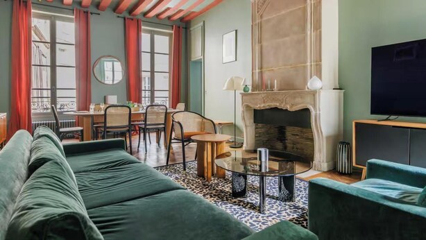 Una sala de estar acogedora con chimenea, sofás verdes, un comedor con sillas de mimbre y un televisor a la derecha.