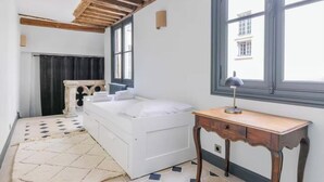 Un dormitorio con una cama blanca, una mesa de madera, un radiador y una ventana con vistas a edificios.
