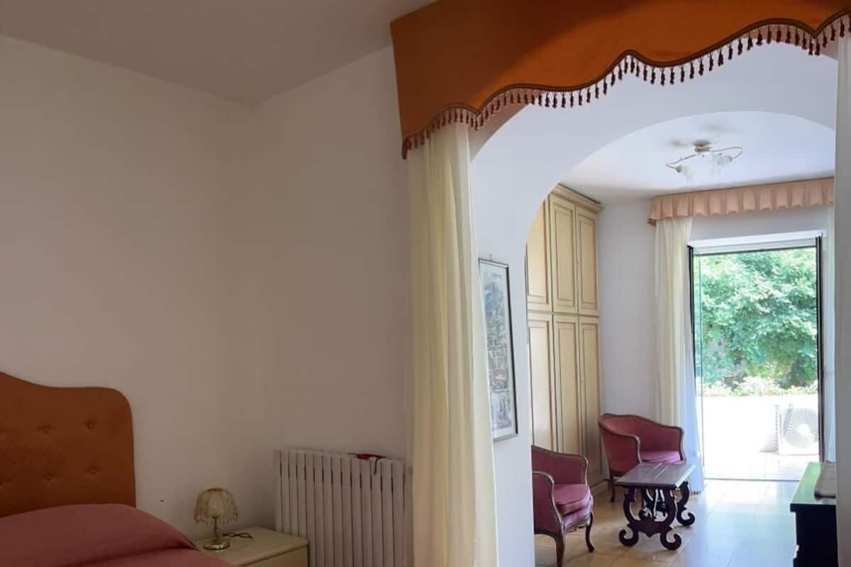 A bedroom with a large bed, a nightstand, a radiator, a chair, a table, and a view of the outdoors.